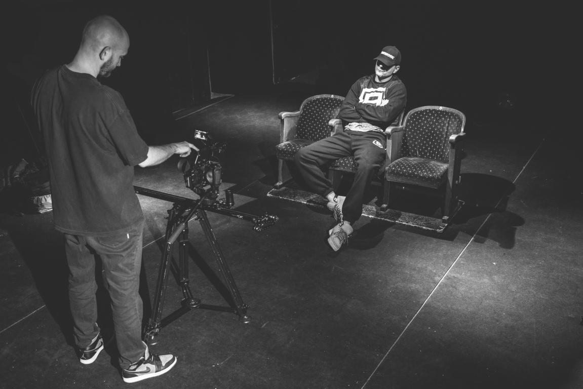 Czarno-białe zdjęcie przedstawia kamerzystę obsługującego profesjonalną kamerę na statywie, skierowaną w stronę młodego mężczyzny siedzącego na krześle w studiu. Mężczyzna w czapce i bluzie ma...