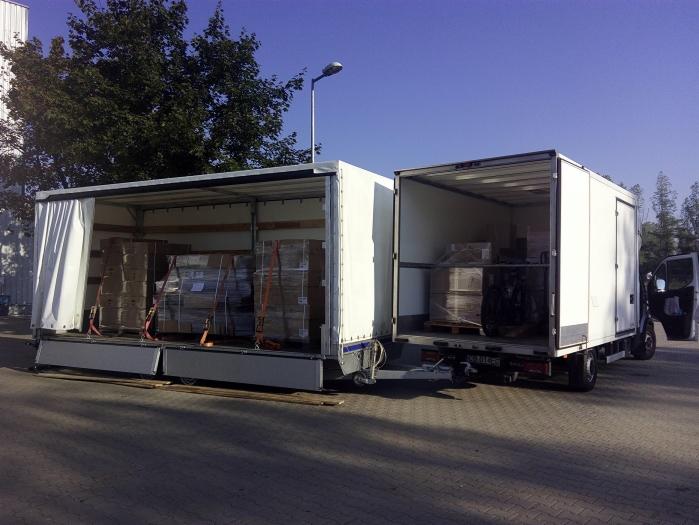 Dwa białe busy z otwartymi przestrzeniami ładunkowymi, załadowane paletami zabezpieczonymi pasami transportowymi, stojące na parkingu w słoneczny dzień.