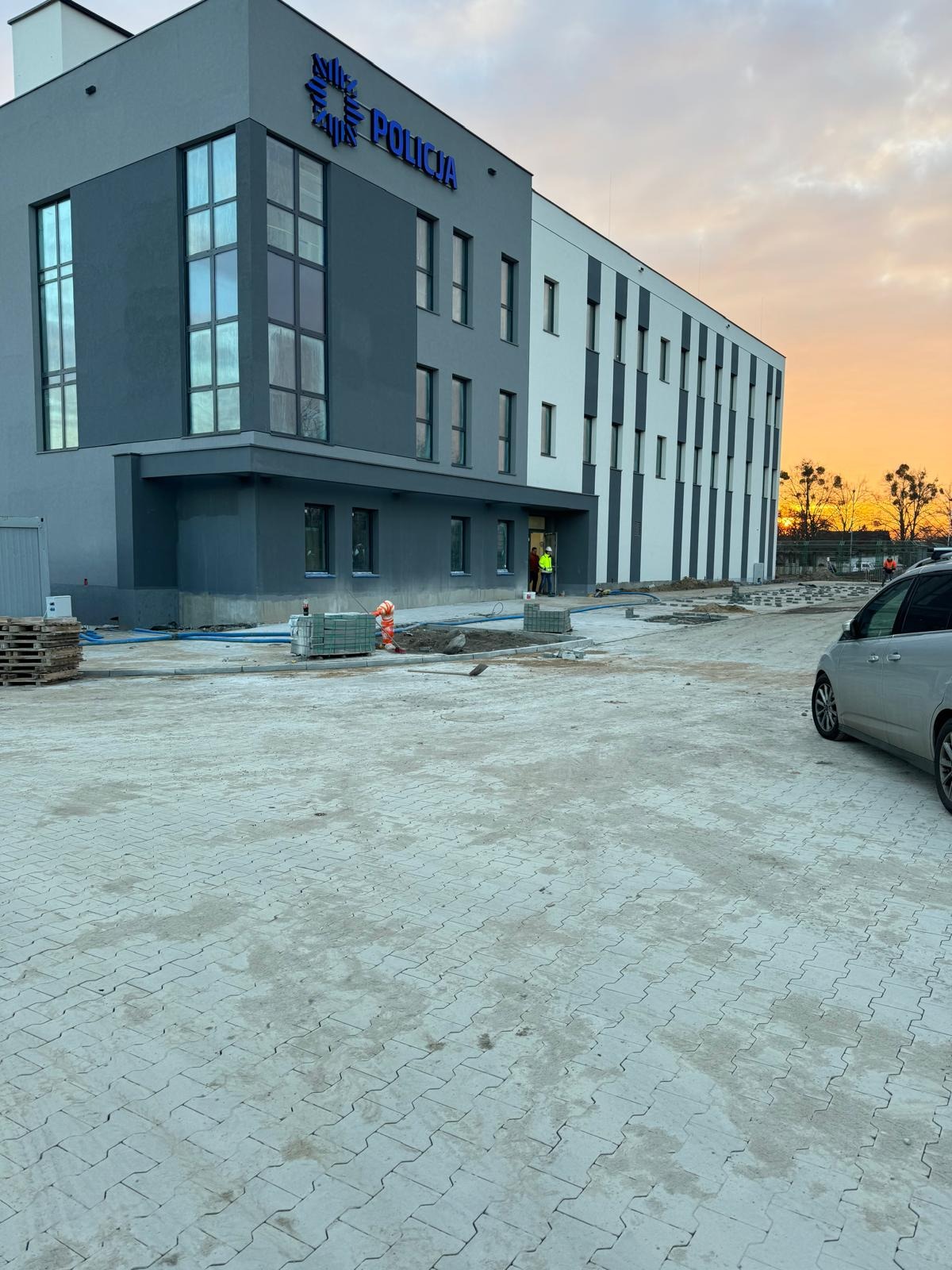 Nowy budynek policji z kostką brukową w trakcie układania, palety z kostką, pomarańczowe pachołki, zachodzące słońce w tle.