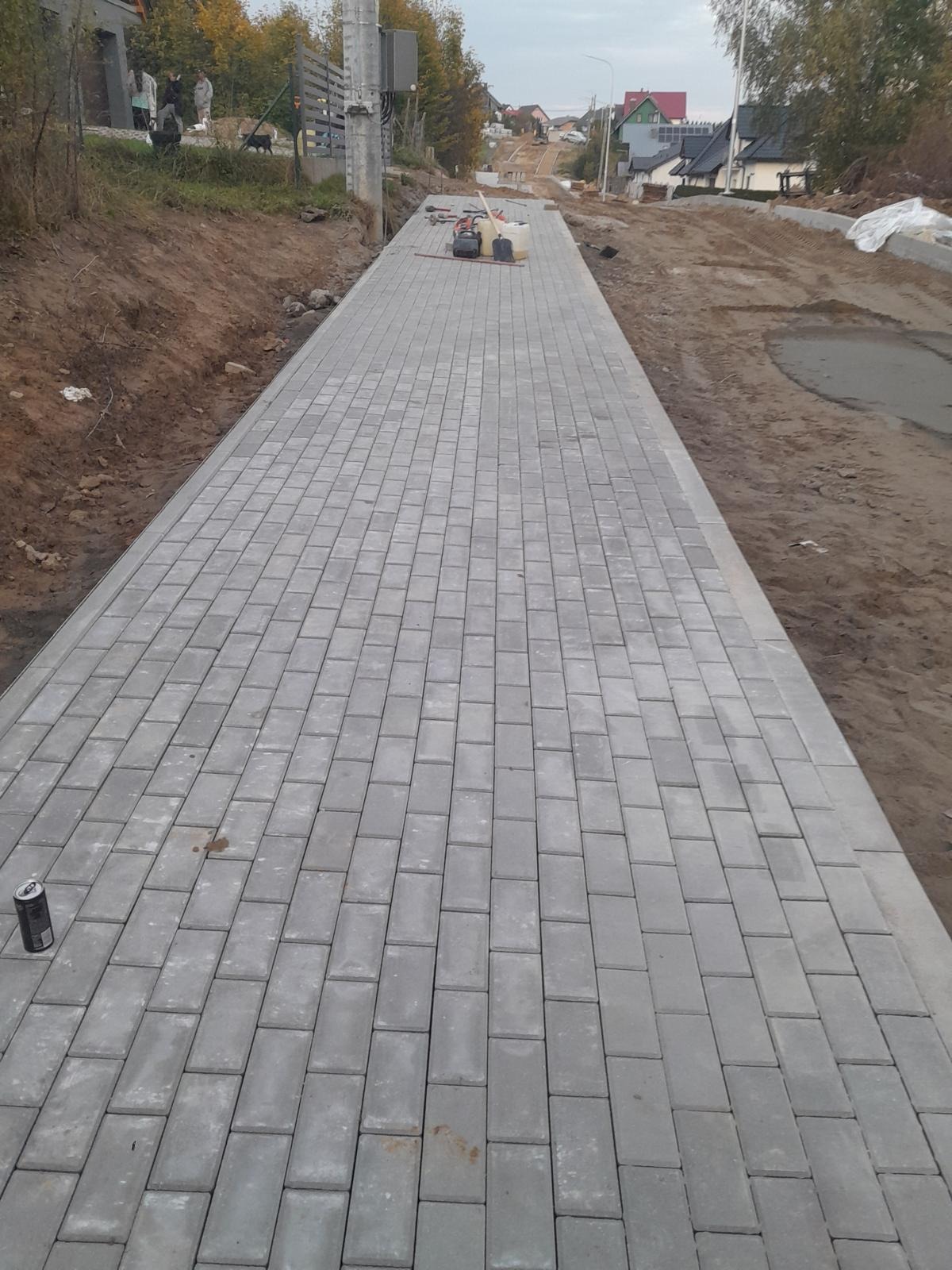 Długa, szara ścieżka z kostki brukowej w trakcie układania, z narzędziami i puszką napoju widocznymi w tle, na tle ziemi i zabudowań.