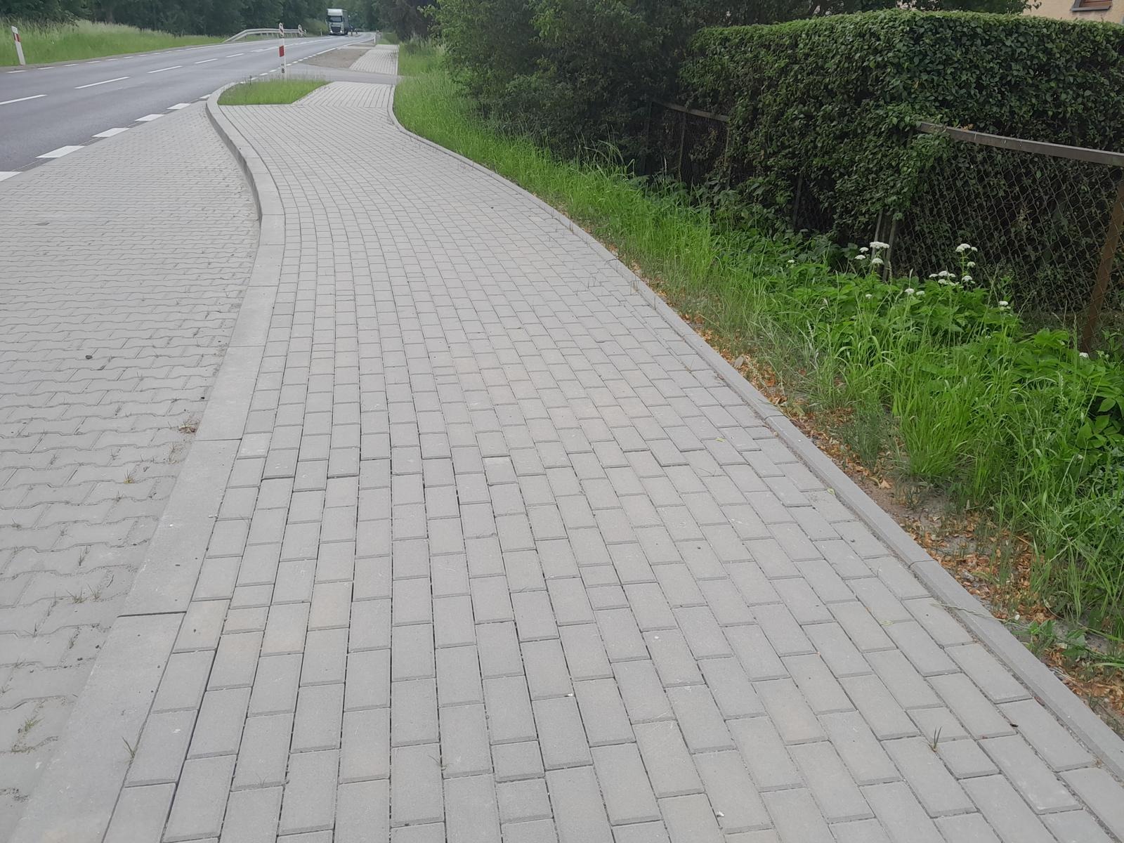 Szara kostka brukowa ułożona wzdłuż drogi, oddzielona obrzeżem, z widoczną zielenią i fragmentem jezdni w tle. Perspektywa z poziomu chodnika.