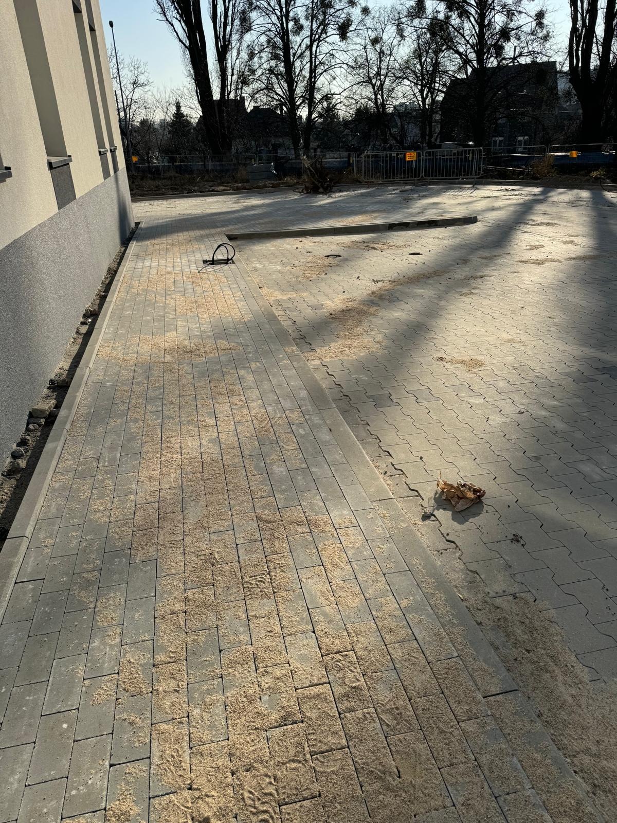 Świeżo ułożona kostka brukowa, zasypana piaskiem, czeka na ostateczne wykończenie. Widoczne ślady butów na piasku i cień drzew na powierzchni.