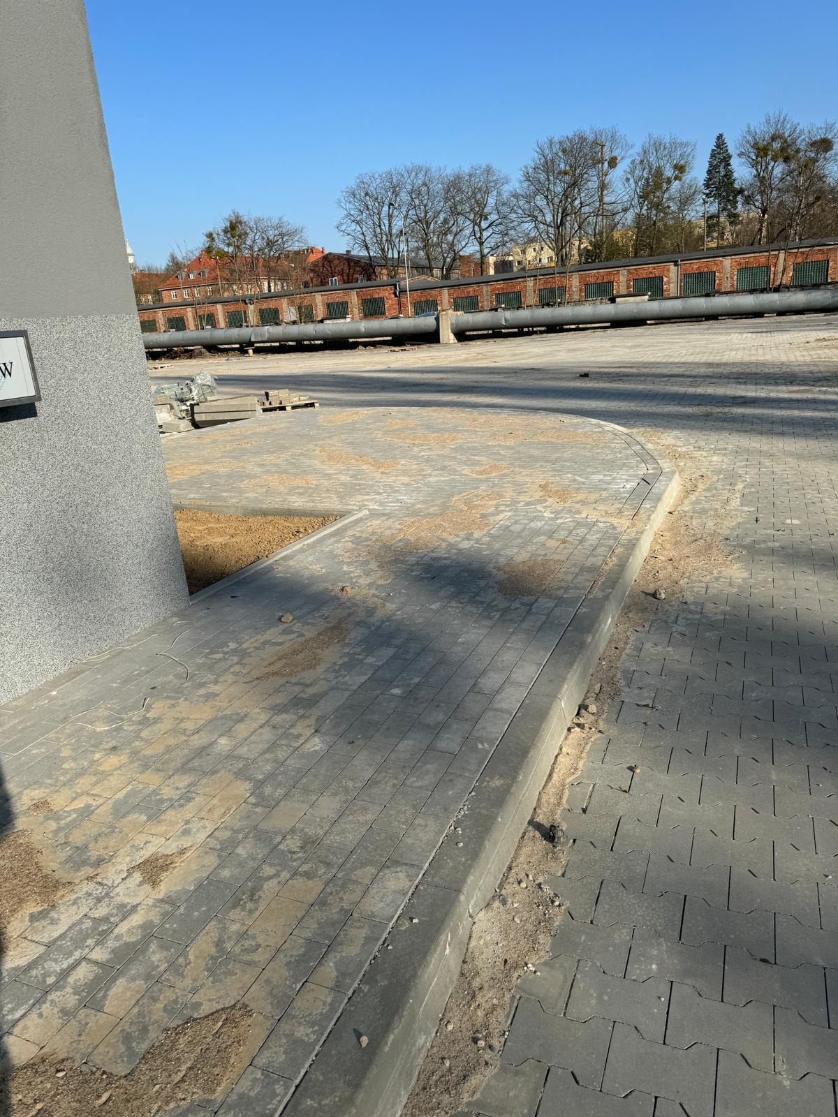 Fragment placu z szarej kostki brukowej, w tle budynek z cegły i rury. Widoczne ślady piasku na kostce, sugerujące prace brukarskie w toku. Słoneczny dzień.