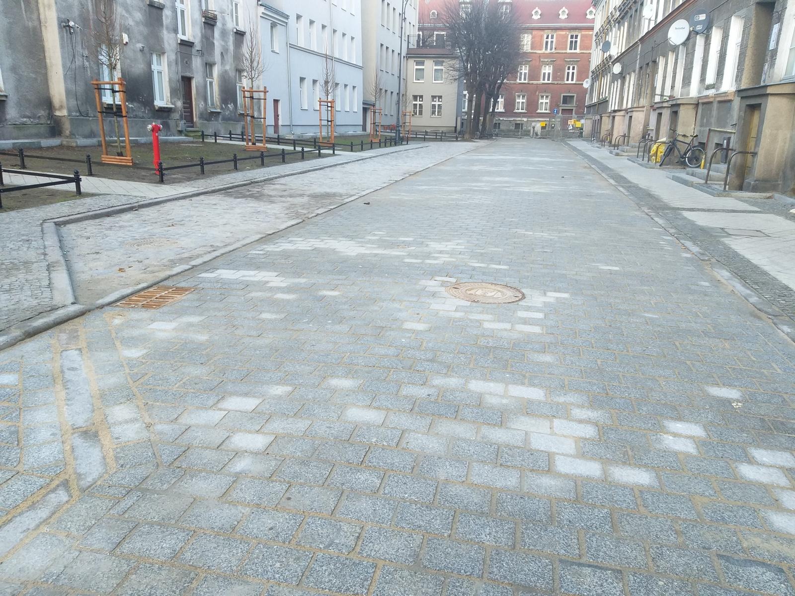 Nowa, jasnoszara kostka granitowa na ulicy miejskiej z hydrantem i młodymi drzewkami. Widok perspektywiczny w kierunku zabudowy.
