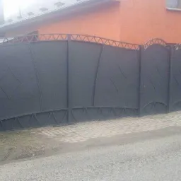 Szara, metalowa brama wjazdowa z dekoracyjnymi elementami w kształcie trójkątów i rombów, na tle pomarańczowego domu i kostki brukowej.