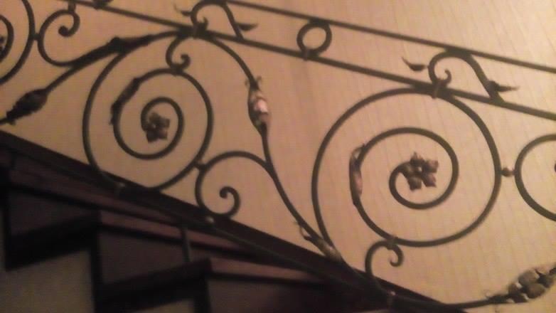 Fragment balustrady schodowej z kutego żelaza, zdobiony motywem roślinnym i spiralnymi zawijasami, widoczny w słabym oświetleniu.