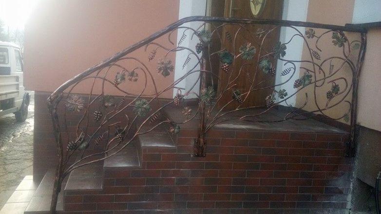 Kute balustrady schodowe z motywem winogron, brązowe, zamontowane na ceglanym podeście, w tle zaparkowany biały samochód dostawczy.