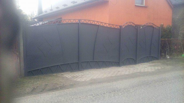 Szara, metalowa brama wjazdowa z dekoracyjnymi elementami w kształcie trójkątów i rombów, na tle pomarańczowego domu i kostki brukowej.