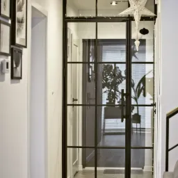 Szklane drzwi wewnętrzne z czarnymi szprosami w korytarzu, widok na salon z roślinami i żaluzjami, dekoracje świąteczne w postaci gwiazdy i bombek.