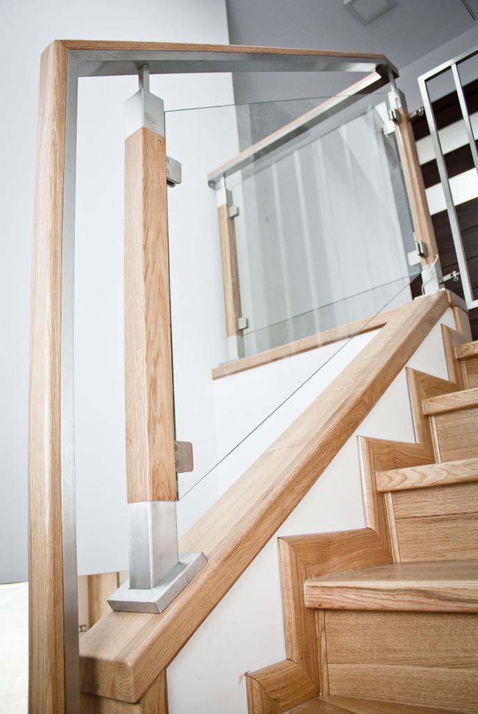 Detale wykończenia schodów drewnianych: połączenie drewna dębowego, stali nierdzewnej i szkła w balustradzie, widoczne mocowania szkła na metalowych elementach, jasne oświetlenie.