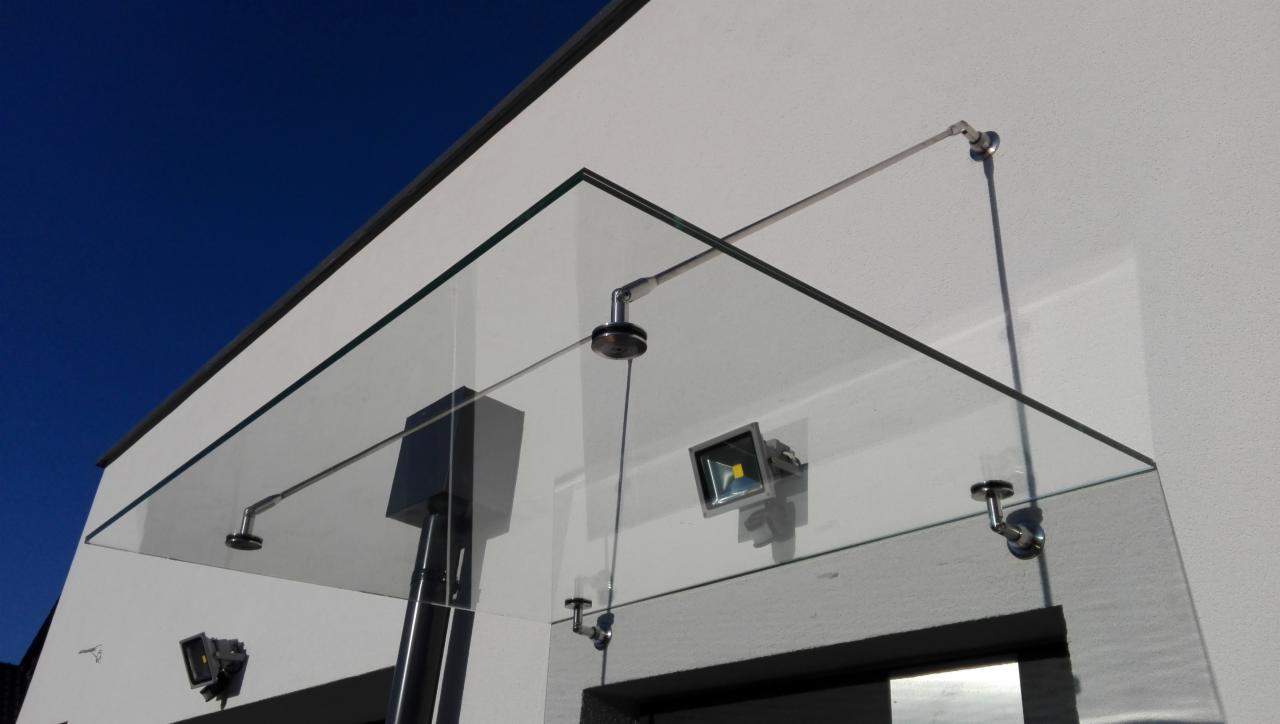 Szklany daszek nad wejściem do budynku, mocowany na metalowych wspornikach, oświetlony punktowymi lampami LED, na tle błękitnego nieba.