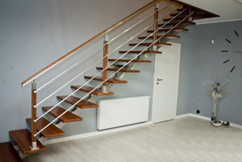 Ażurowe schody z drewnianymi stopniami i balustradą z drewnianą poręczą i metalowymi wypełnieniami, widok z dołu na tle szarej ściany z dekoracyjnym zegarem i białą grzejnikiem.