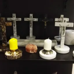 Ekspozycja nagrobków krzyży z marmuru i granitu na czarnej półce, obok świeczniki z marmurowymi podstawami i kolorowymi świecami oraz bryła soli kamiennej.