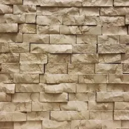 Ściana wyłożona trójwymiarowymi, beżowymi płytkami z kamienia naturalnego o nieregularnej fakturze, ułożonymi w prostokątny wzór.