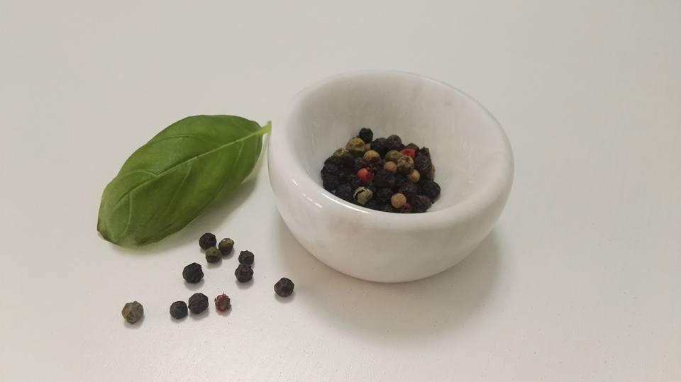 Mała, biała miseczka z marmuru wypełniona mieszanką kolorowego pieprzu, obok liść bazylii i kilka ziaren pieprzu na białym tle.
