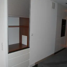 Nowoczesna zabudowa kuchenna z roletą, drewnianymi półkami i białymi szufladami, w minimalistycznym wnętrzu.