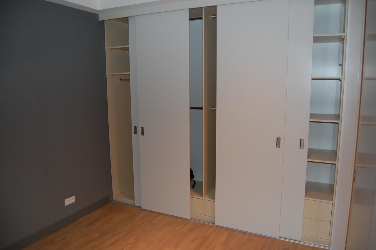 Wnętrze pokoju z zabudowaną szafą z przesuwnymi, białymi drzwiami. Widoczne półki i drążki na ubrania. Ściana w kolorze ciemnoszarym, podłoga drewniana.