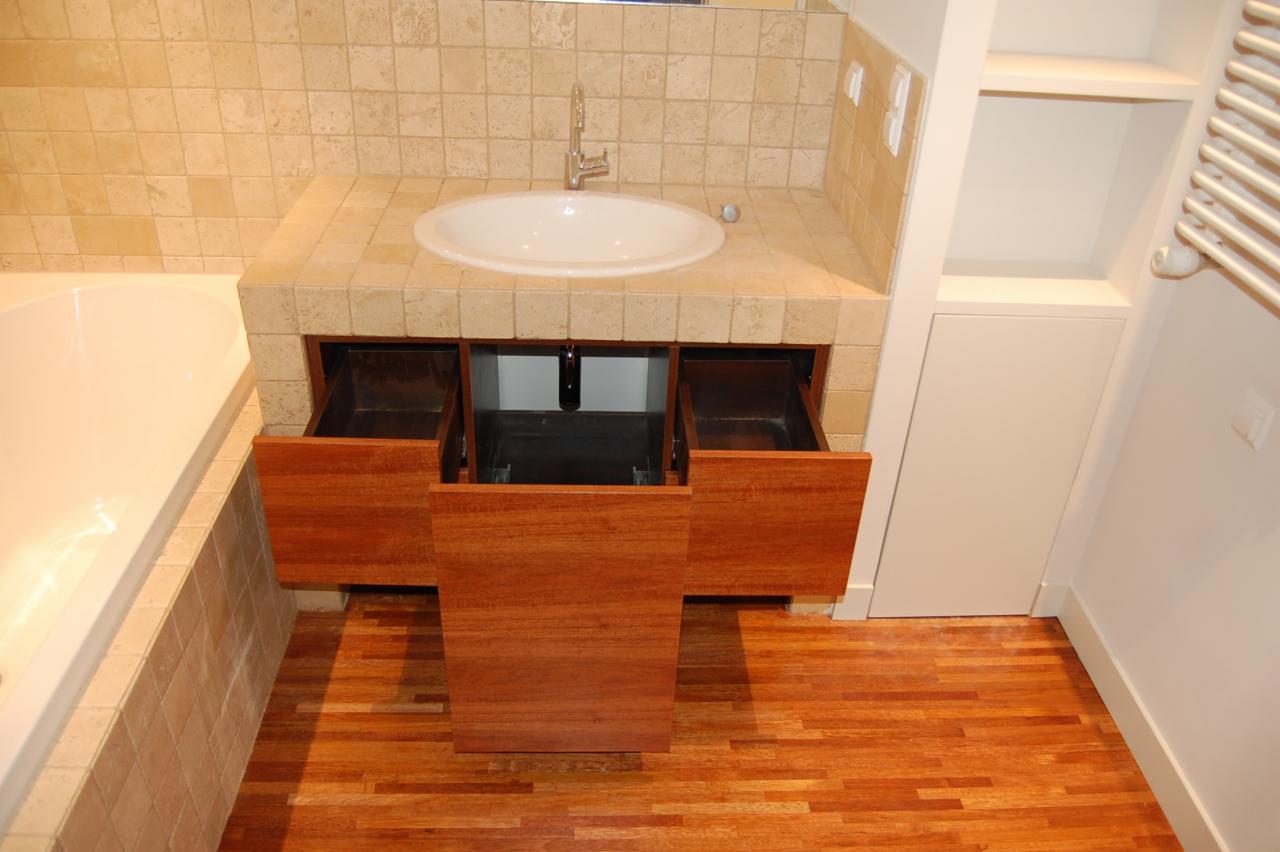 Łazienka z umywalką wpuszczoną w kamienny blat, szafka z drewna egzotycznego z otwartymi szufladami, parkiet z drobnych klepek, beżowe płytki na ścianach, obok wanna i zabudowa z półkami.