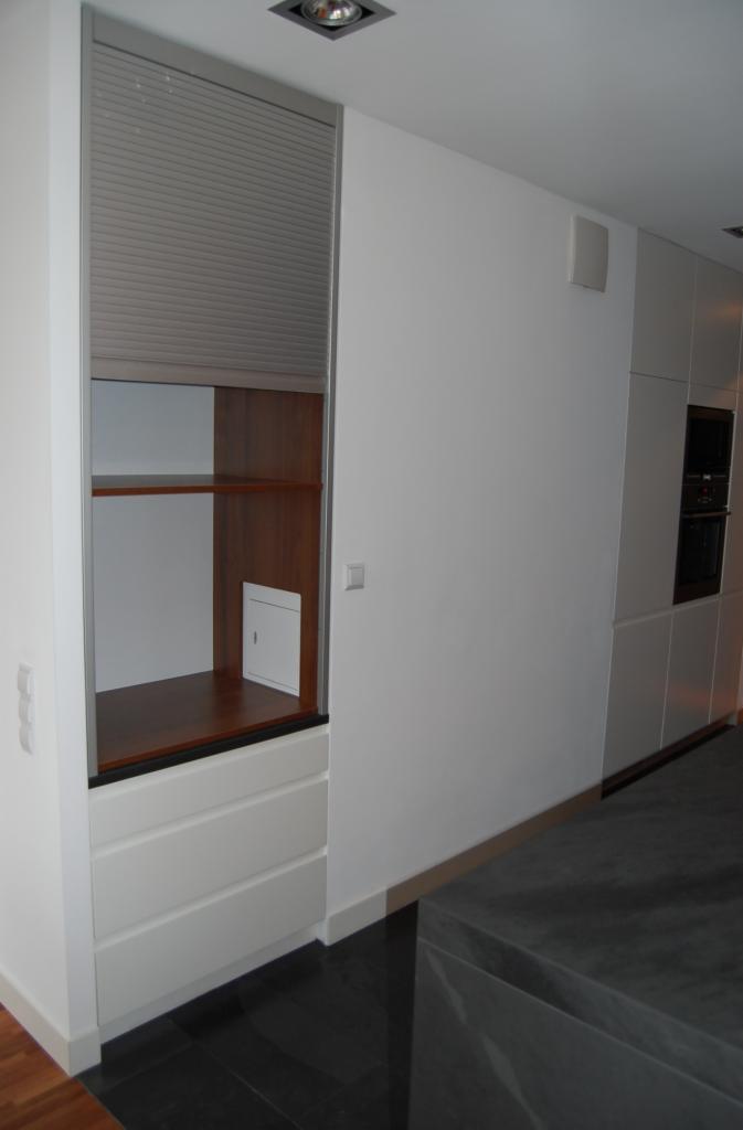 Nowoczesna zabudowa kuchenna z roletą, drewnianymi półkami i białymi szufladami, w minimalistycznym wnętrzu.