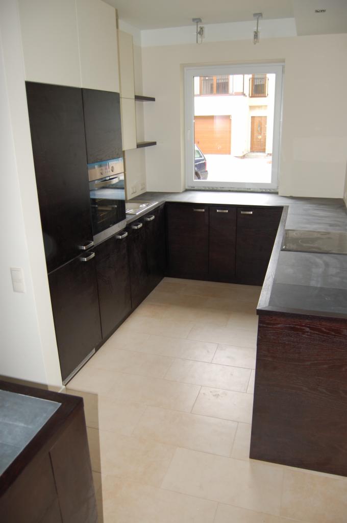 Nowoczesna kuchnia z ciemnobrązowymi, drewnianymi szafkami z metalowymi uchwytami i jasnym blatem, widok na okno z zabudową zewnętrzną.