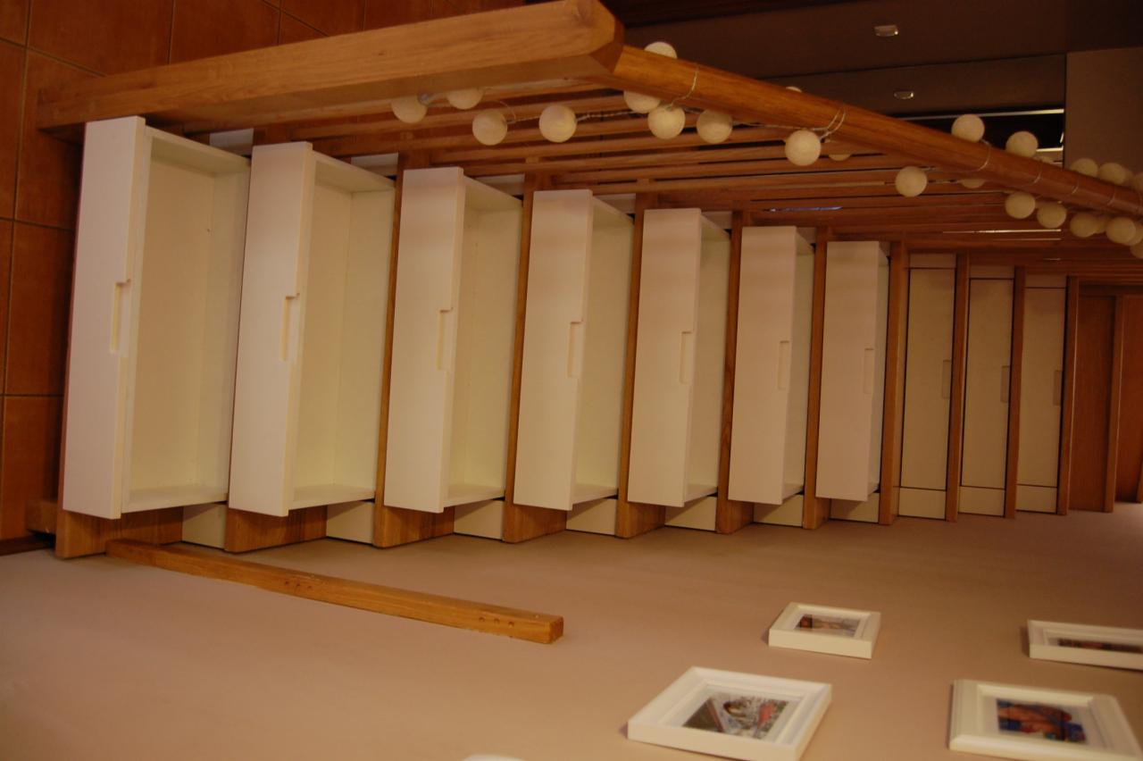 Nietypowy widok na białe, otwarte szafki z drewnianymi elementami konstrukcyjnymi, oświetlone sznurem kulistych lamp, z ramkami zdjęć na podłodze.