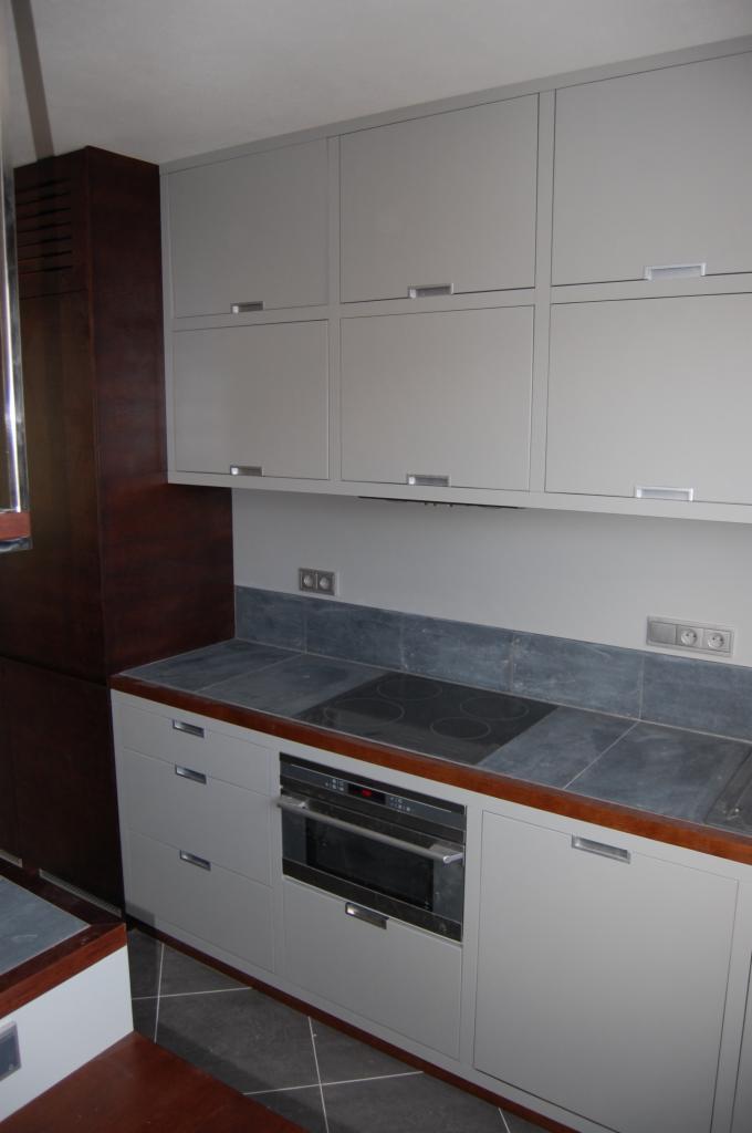 Szare szafki kuchenne z ciemnym blatem, wbudowaną płytą grzewczą i piekarnikiem, widoczne gniazdka elektryczne na ścianie.