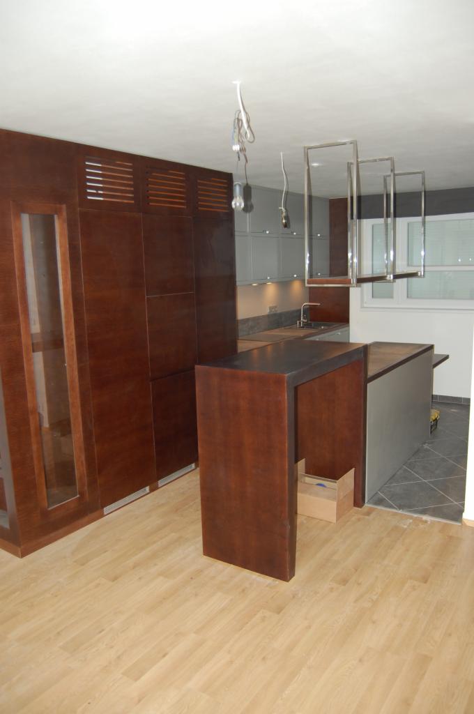 Nowoczesna kuchnia z drewnianą zabudową, wyspą i oświetleniem wiszącym, widok na szare szafki i okno.