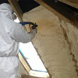 Pracownik w białym kombinezonie ochronnym natryskuje pianę izolacyjną na skośny dach poddasza, widoczne krople piany w powietrzu.