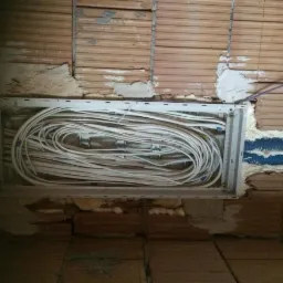 Skrzynka z dużą ilością białych kabli ułożonych w pętle, zamontowana w surowej, ceglanej ścianie z widocznymi śladami zaprawy i pianki montażowej.