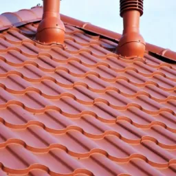 Czerwona blacha dachowa z dwoma odpowietrznikami kominowymi na tle jasnego nieba, widok z dołu.