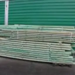 Stos impregnowanych desek na palecie, widoczny na tle zielonej ściany magazynu.