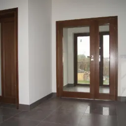 Dwa rodzaje brązowych drzwi z przeszkleniami: jedne wewnętrzne z matowym szkłem, drugie zewnętrzne przesuwne z widokiem na ogród, na tle białych ścian i szarej podłogi.