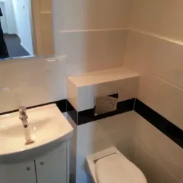 Nowoczesna łazienka z białymi płytkami, czarnymi akcentami, wiszącą toaletą i umywalką z szafką, widoczny chromowany przycisk spłuczki i lustro.