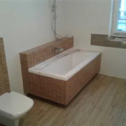 Łazienka po remoncie: zabudowana wanna wykończona mozaiką drewnopodobną, toaleta z podtynkową spłuczką również w mozaice, płytki podłogowe imitujące drewno.