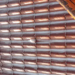 Nowa, błyszcząca dachówka ceramiczna w kolorze miedzianym, częściowo pokryta białą warstwą ochronną, widoczna z perspektywy rusztowania z pomarańczowych rur.