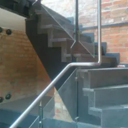 Nowoczesne schody z ciemnego kamienia z balustradą ze stali nierdzewnej i szkła, na tle ceglanej ściany z widocznymi śrubami mocującymi.