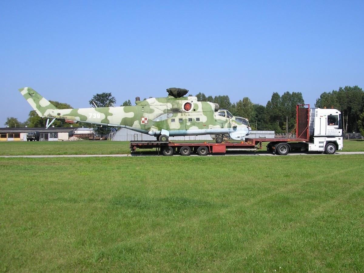 Kamuflażowany helikopter z numerem 730 przewożony na niskopodwoziowej platformie przez białą ciężarówkę na tle zielonego trawnika i budynków.