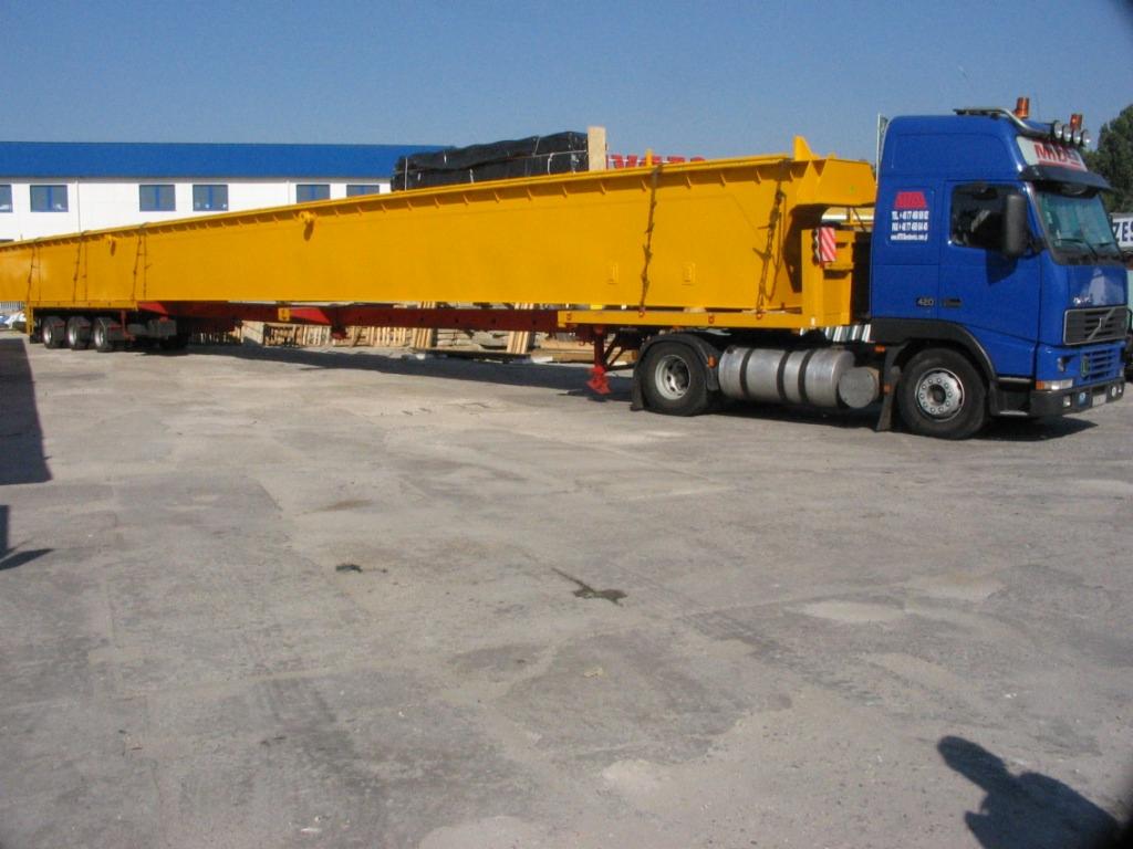 Niebieska ciężarówka Volvo z żółtą, długą i szeroką konstrukcją stalową na naczepie, zaparkowana na betonowym placu, z budynkiem w tle.