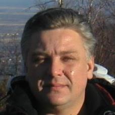 Portret mężczyzny z siwiejącymi włosami na tle rozmytego krajobrazu miejskiego i drzew bez liści.