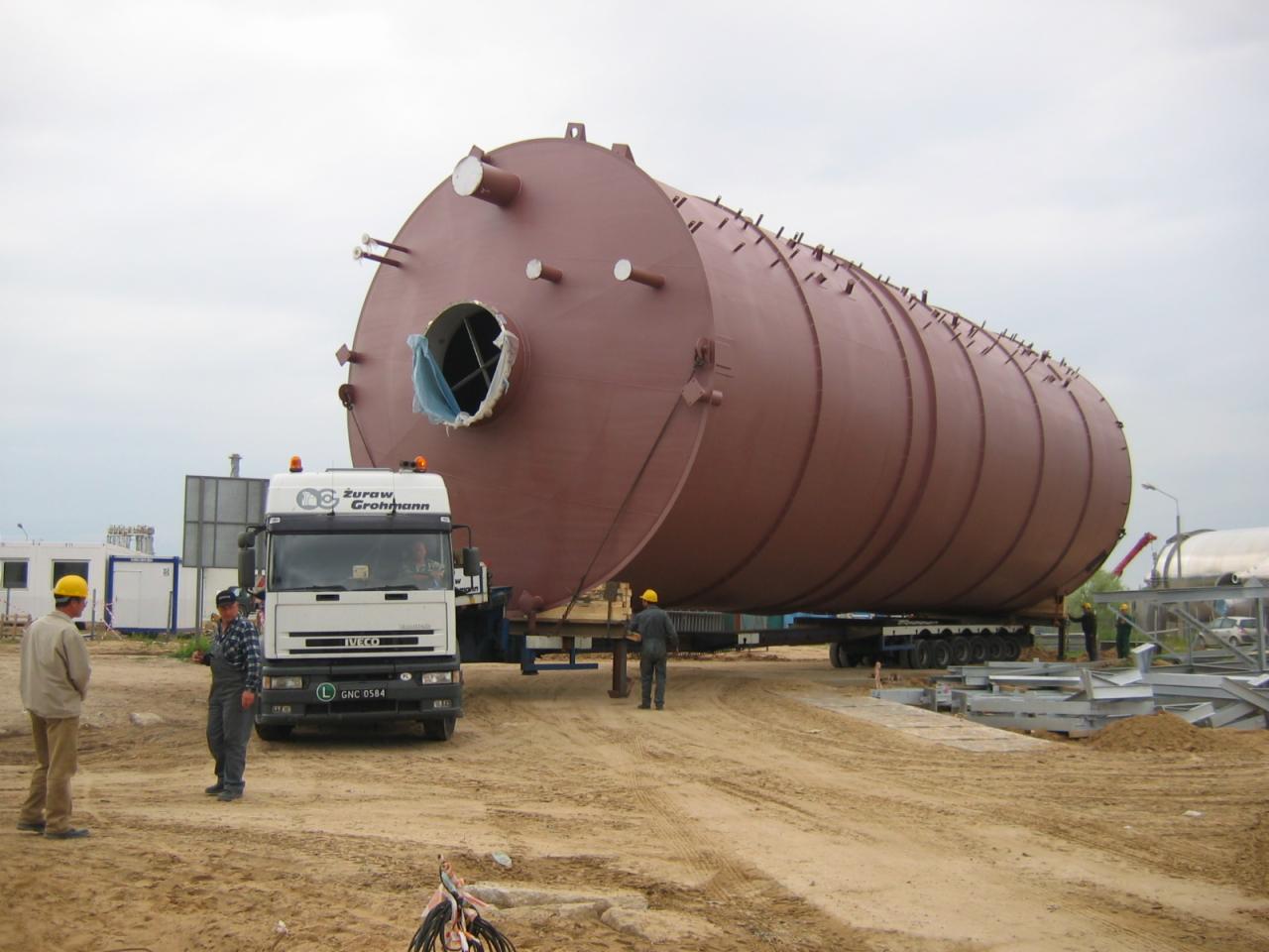 Transport specjalistyczny: Duży, brązowy zbiornik przemysłowy przewożony na naczepie niskopodwoziowej ciężarówką Iveco, z widocznymi pracownikami w kaskach i elementami placu budowy w tle.