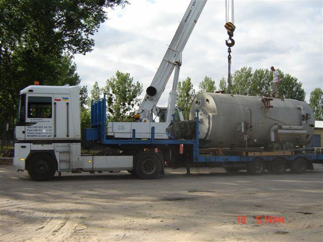 W Białymstoku dźwig umieszcza duży, szary zbiornik przemysłowy na platformie ciężarówki. Na zbiorniku stoi mężczyzna, a w tle widać drzewa.