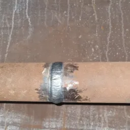 Spoina na rurze z widocznymi śladami spawania i zmatowieniami, na tle metalowej powierzchni z zaciekami. Prezentacja umiejętności spawacza.