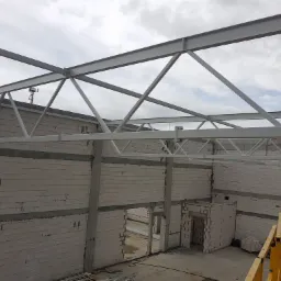 Wnętrze hali w trakcie budowy z widoczną stalową konstrukcją dachu i ścianami z bloczków betonowych, w tle widoczny dźwig budowlany.