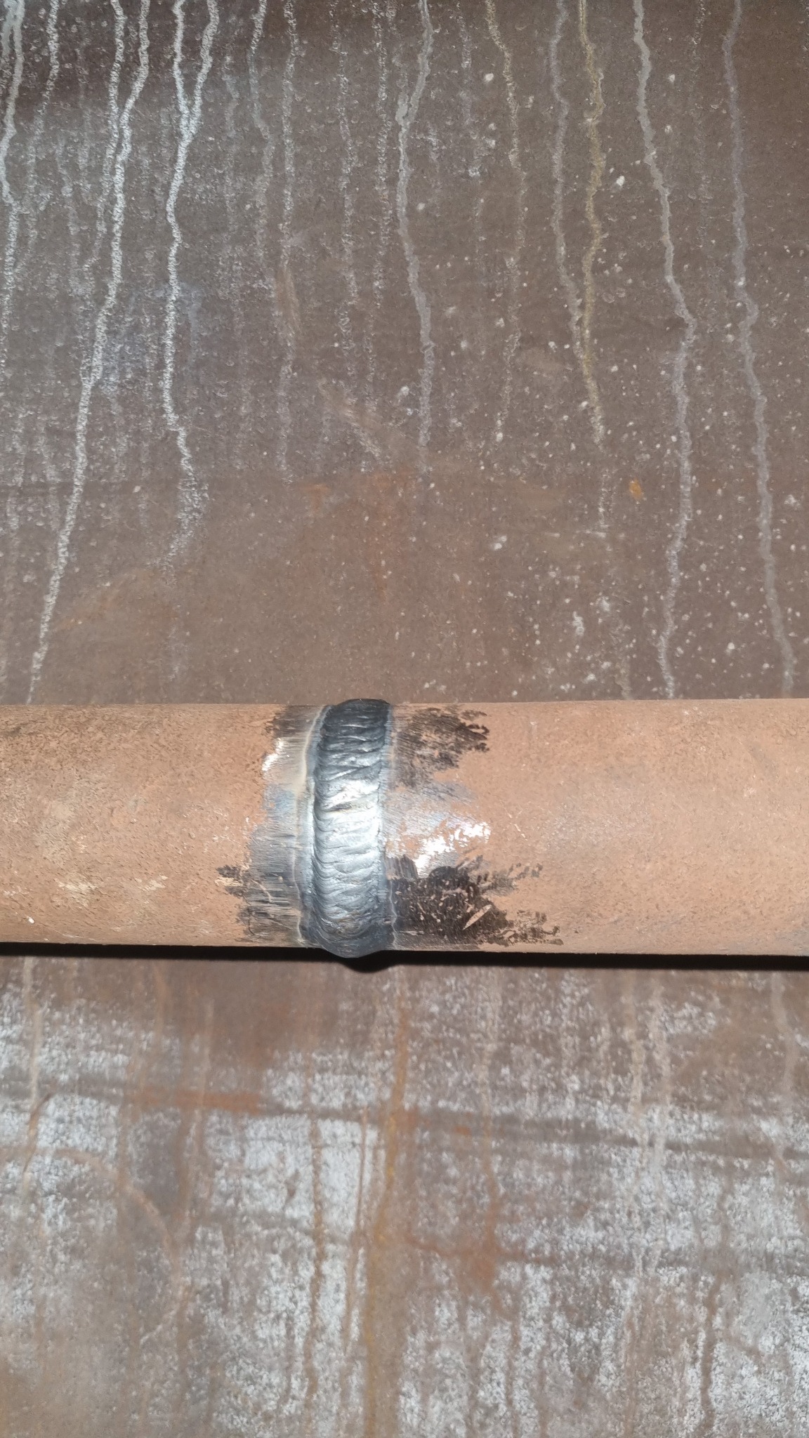 Spoina na rurze z widocznymi śladami spawania i zmatowieniami, na tle metalowej powierzchni z zaciekami. Prezentacja umiejętności spawacza.