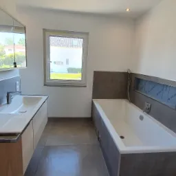 Jasne wnętrze łazienki z nowoczesną białą wanną i umywalką z szafką z jasnego drewna, szare płytki częściowo na ścianach, okno w tle, widoczne gniazdka elektryczne.