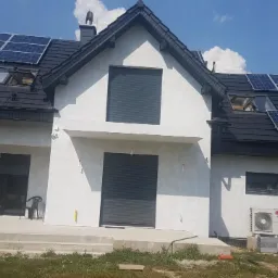 Nowoczesny dom jednorodzinny z szarą elewacją, panelami słonecznymi na czarnym dachu, otwartym oknem dachowym, zewnętrzną jednostką klimatyzacji LG i częściowo opuszczonymi roletami zewnętrznymi.