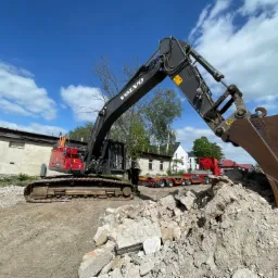Ciężka koparka Volvo na gąsienicach z operatorem w kombinezonie obok, wysypująca gruz na placu budowy, w tle budynki i ciężarówka z naczepą, błękitne niebo z chmurami.