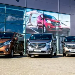 Trzy busy Opel Vivaro w różnych kolorach (brązowy, szary, beżowy) zaparkowane przed salonem samochodowym z dużymi oknami, na których widnieje grafika przedstawiająca czerwony bus.