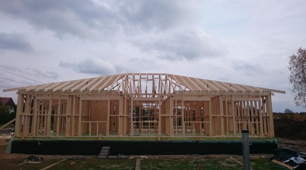 Wznoszenie konstrukcji domu z drewnianych belek na przygotowanym fundamencie, widoczne elementy szkieletu ścian i dachu na tle pochmurnego nieba.