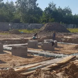 Wykopane rowy pod fundamenty z ułożonymi bloczkami betonowymi, dwóch pracowników w tle, przygotowanie do wylewania betonu.